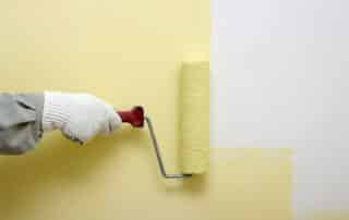 Painter in white gloves applying pale yellow paint to drywall with foam roller How Many Coats of Paint